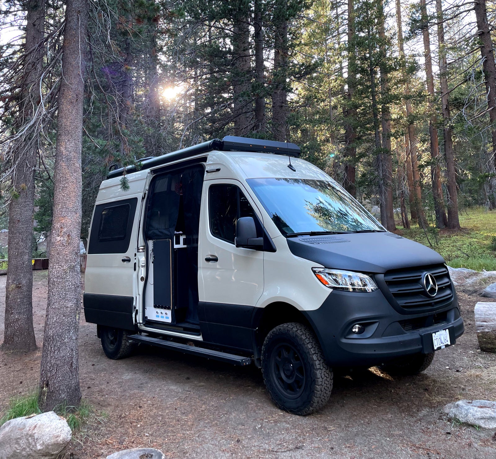 A two-tone Mercedes Sprinter camper van, likely an RV rental, is parked amidst tall trees in a sunny forest.