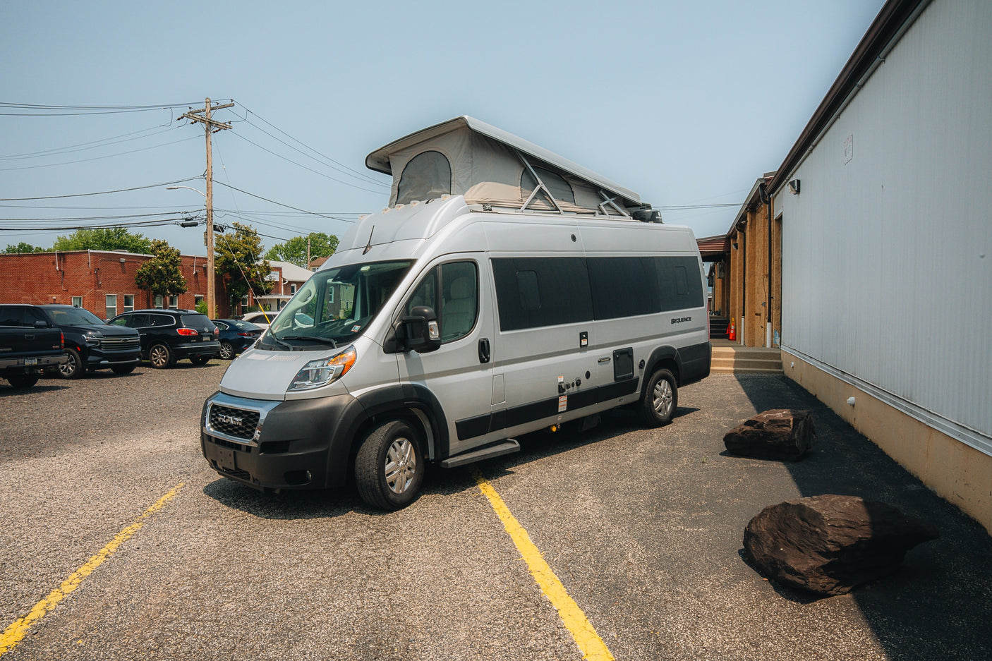 A sleek silver pop-top camper van, an ideal RV rental, parked on a gravel lot with other vehicles and buildings.