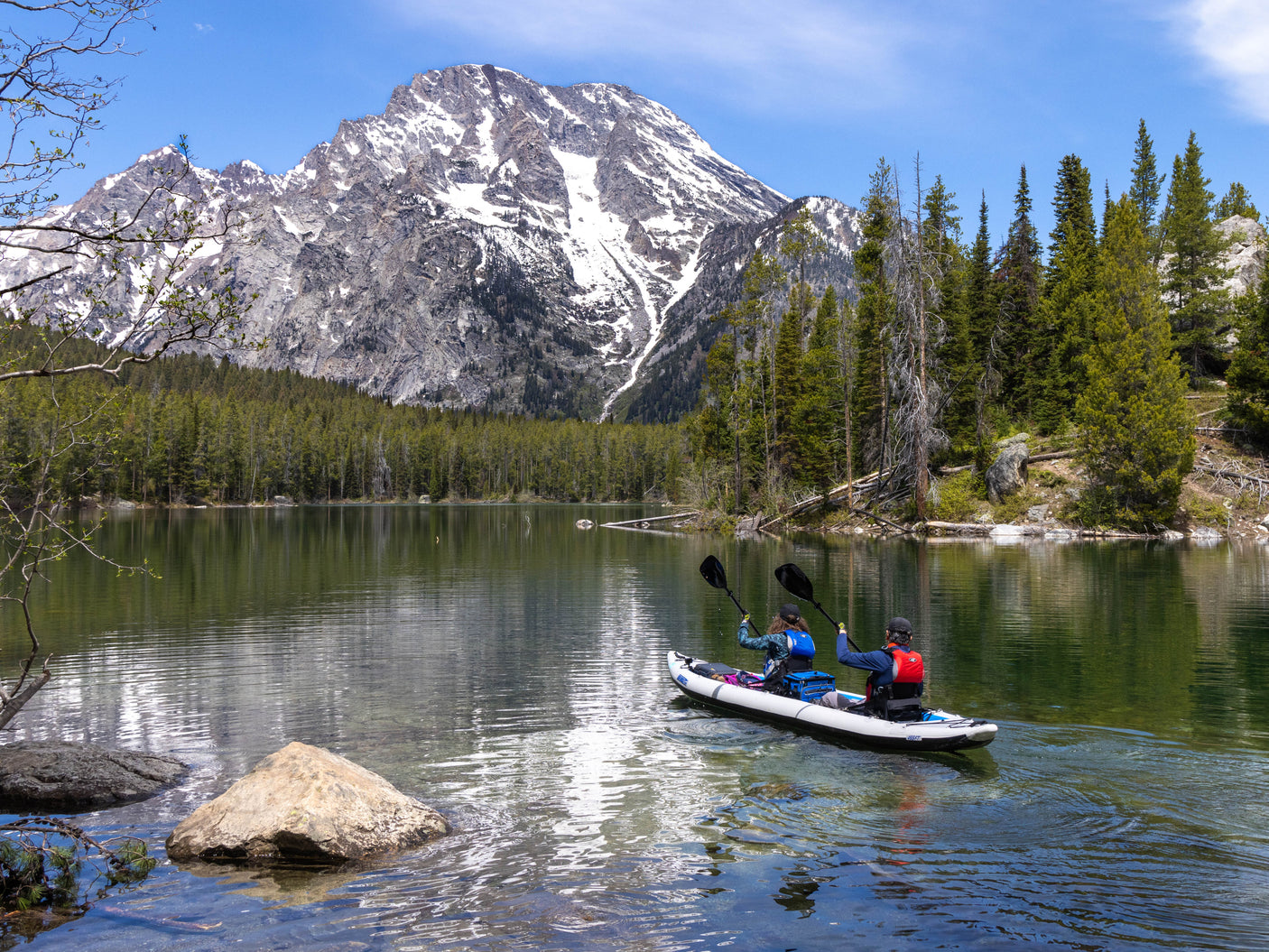Two people paddle their kayak on a clear mountain lake, framed by pine forests and snowy peaks – ideal for an RV rental trip.