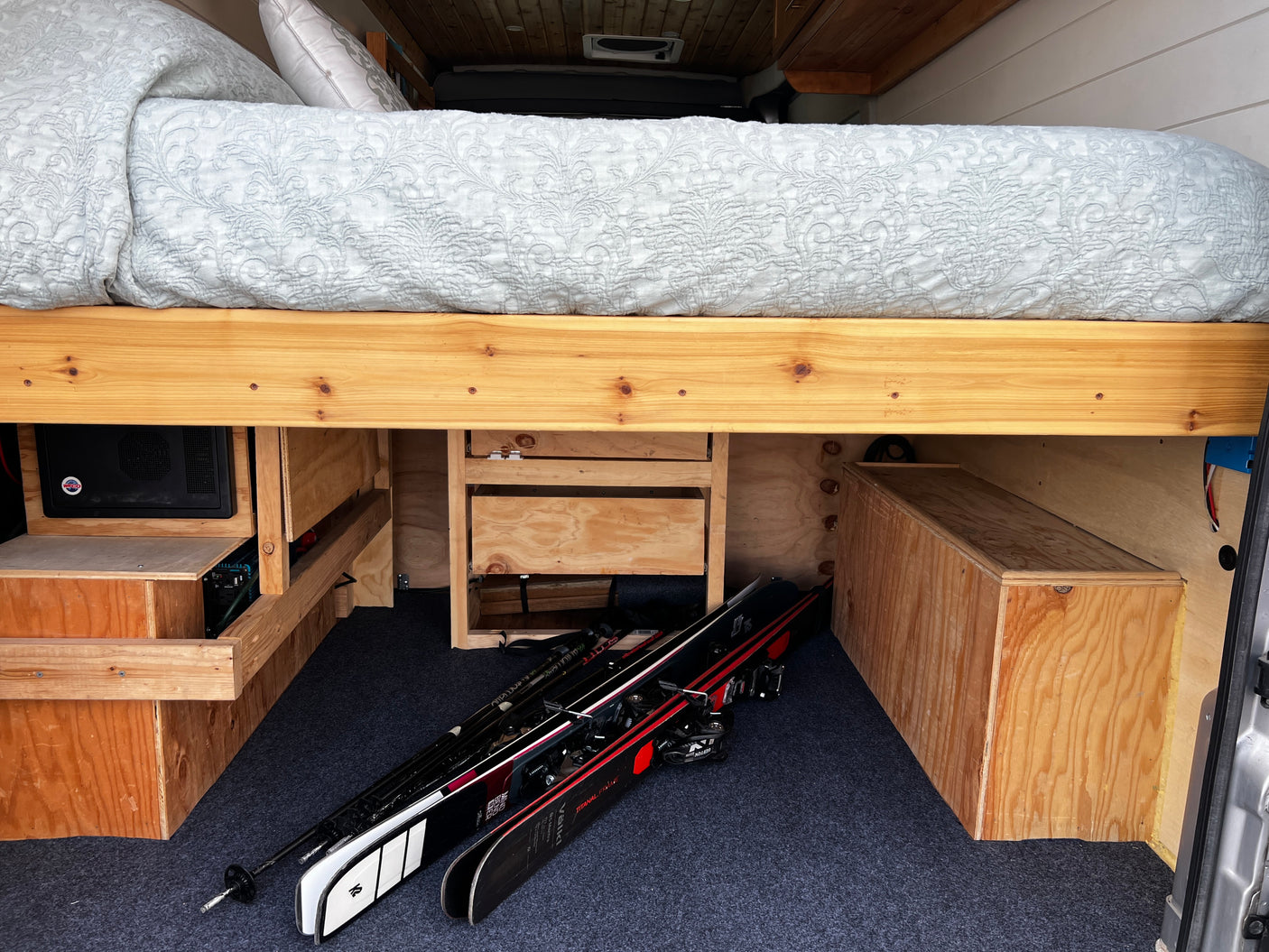 RV rental interior with an elevated bed, custom storage compartments, and skis on the carpeted floor, ready for adventure.