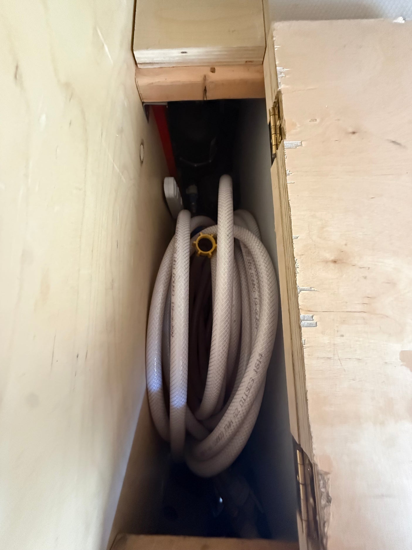RV rental: Coiled white water hose and plumbing connections visible in a narrow wooden storage compartment.