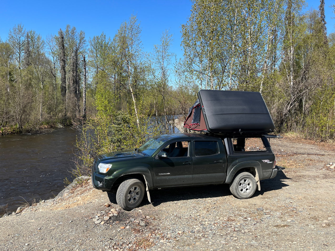 Dark green Tacoma truck with an open roof-top tent beside a river, showcasing an ideal RV rental for outdoor adventures.