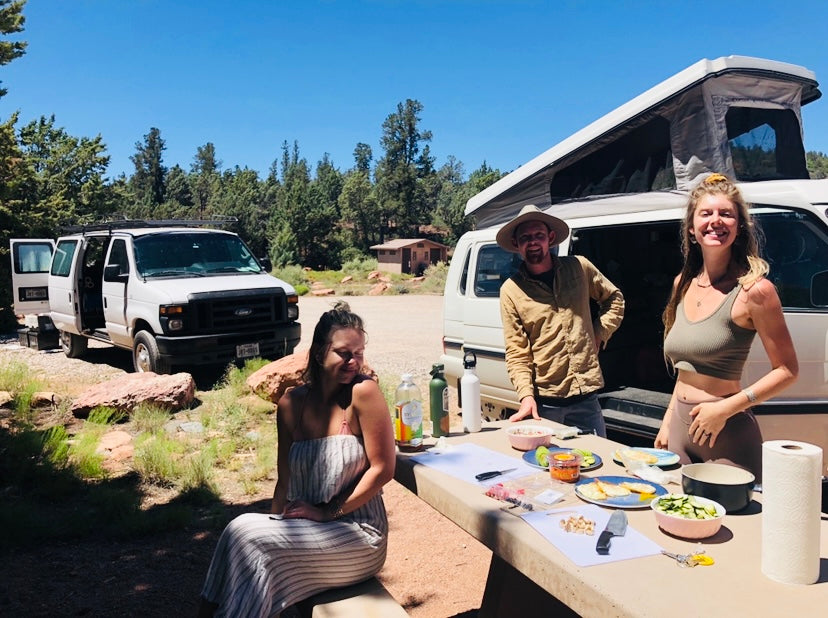 Three friends enjoy meal prep at a sunny campsite with an RV rental and camper vans, embracing outdoor adventures.