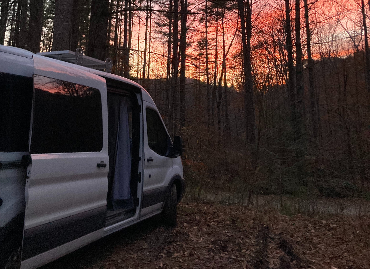 White camper van (rv rental) parked in a rustic forest during a vibrant orange and pink sunset, ideal for nature escapes.