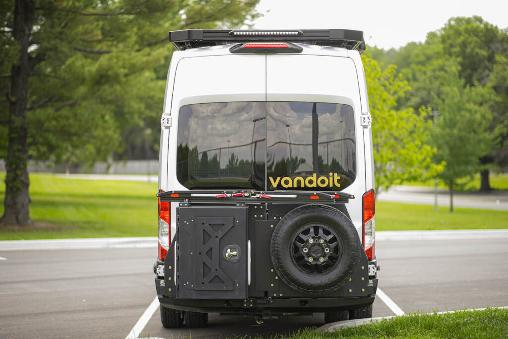 Rear view of a white custom camper van for RV rental, with a spare tire and storage on the back, parked outdoors.