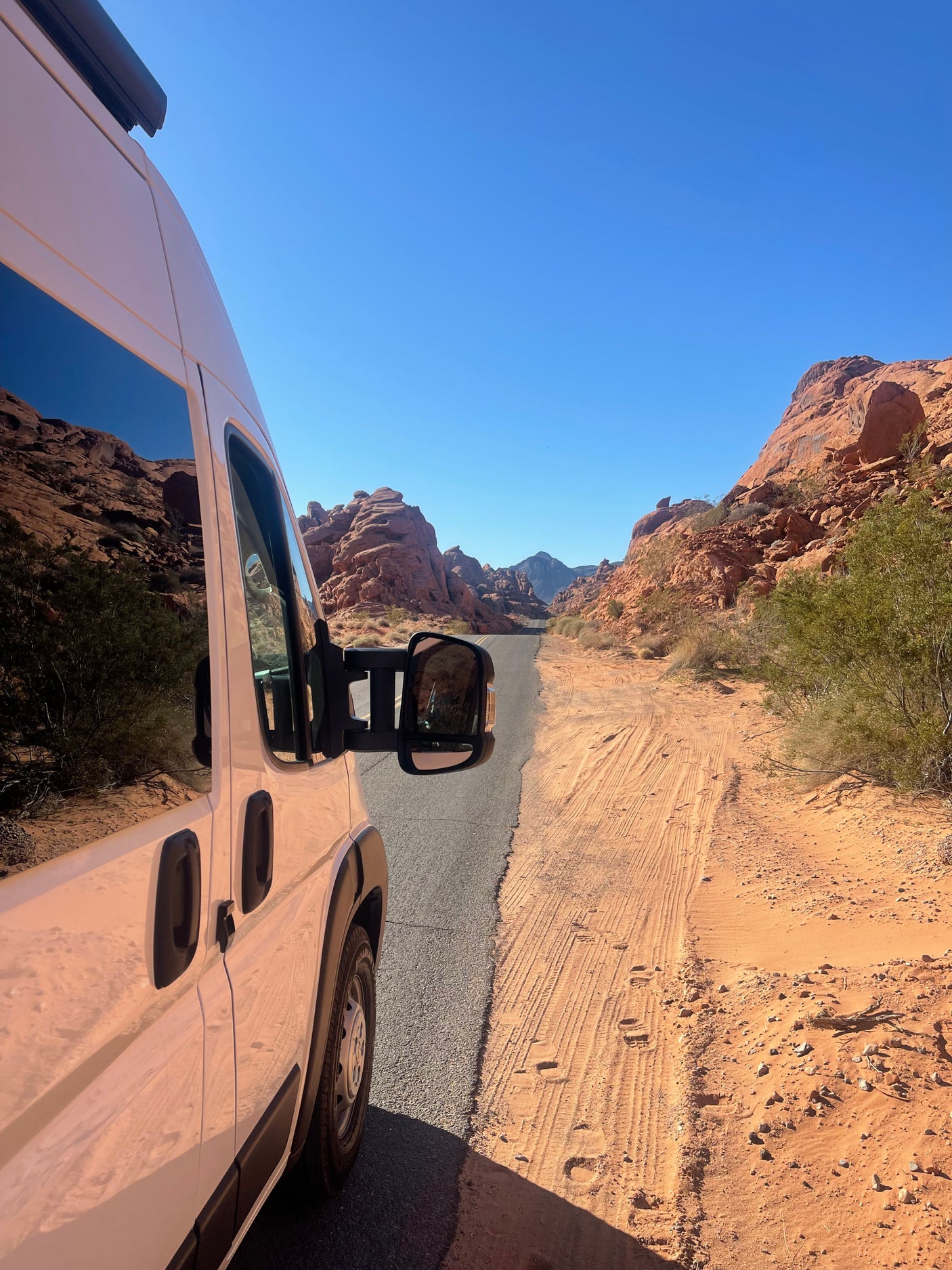 White RV rental on a desert road, flanked by towering red rock formations under a clear blue sky.