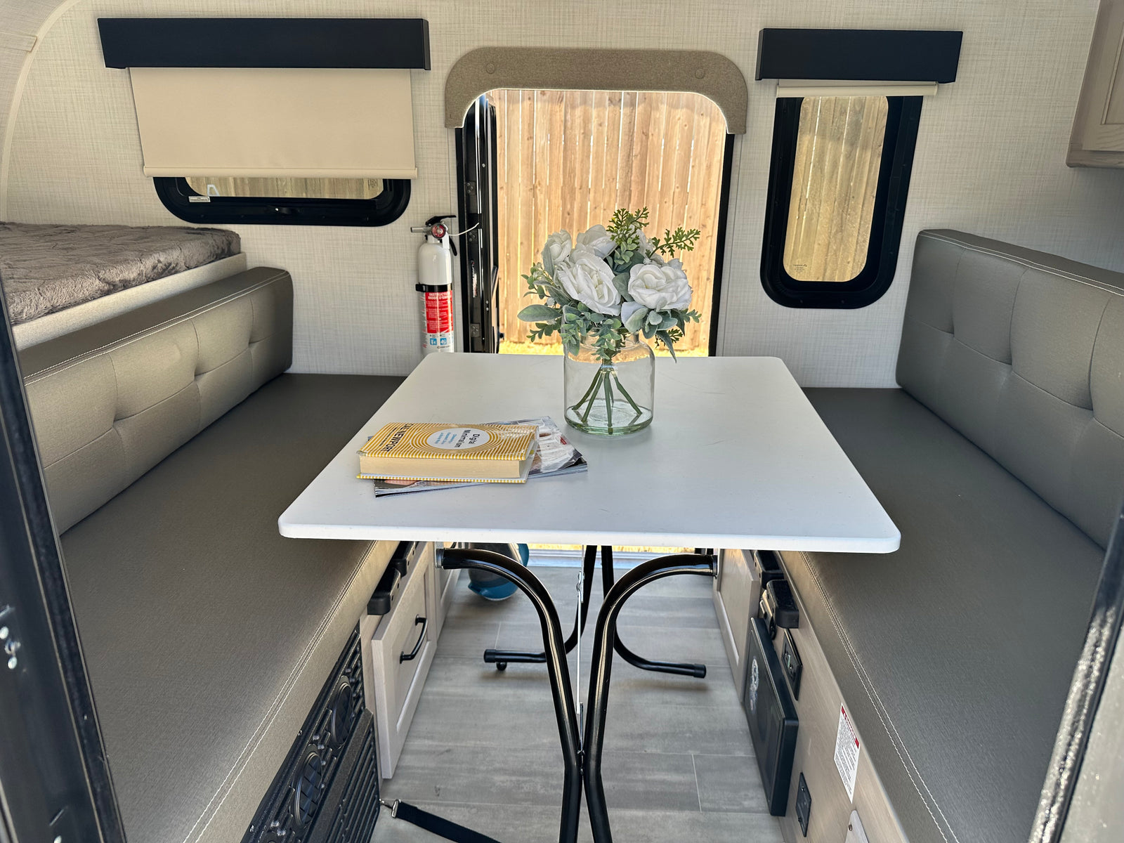 Bright RV rental interior features a cozy dinette with a white table, flowers, a book, and a comfy bed. Ready for adventure.