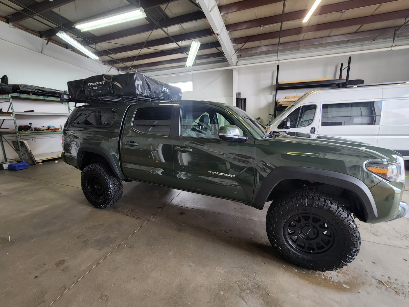 Green Tacoma with rooftop tent & white RV rental van in a workshop. Off-road camping setup, adventure ready.
