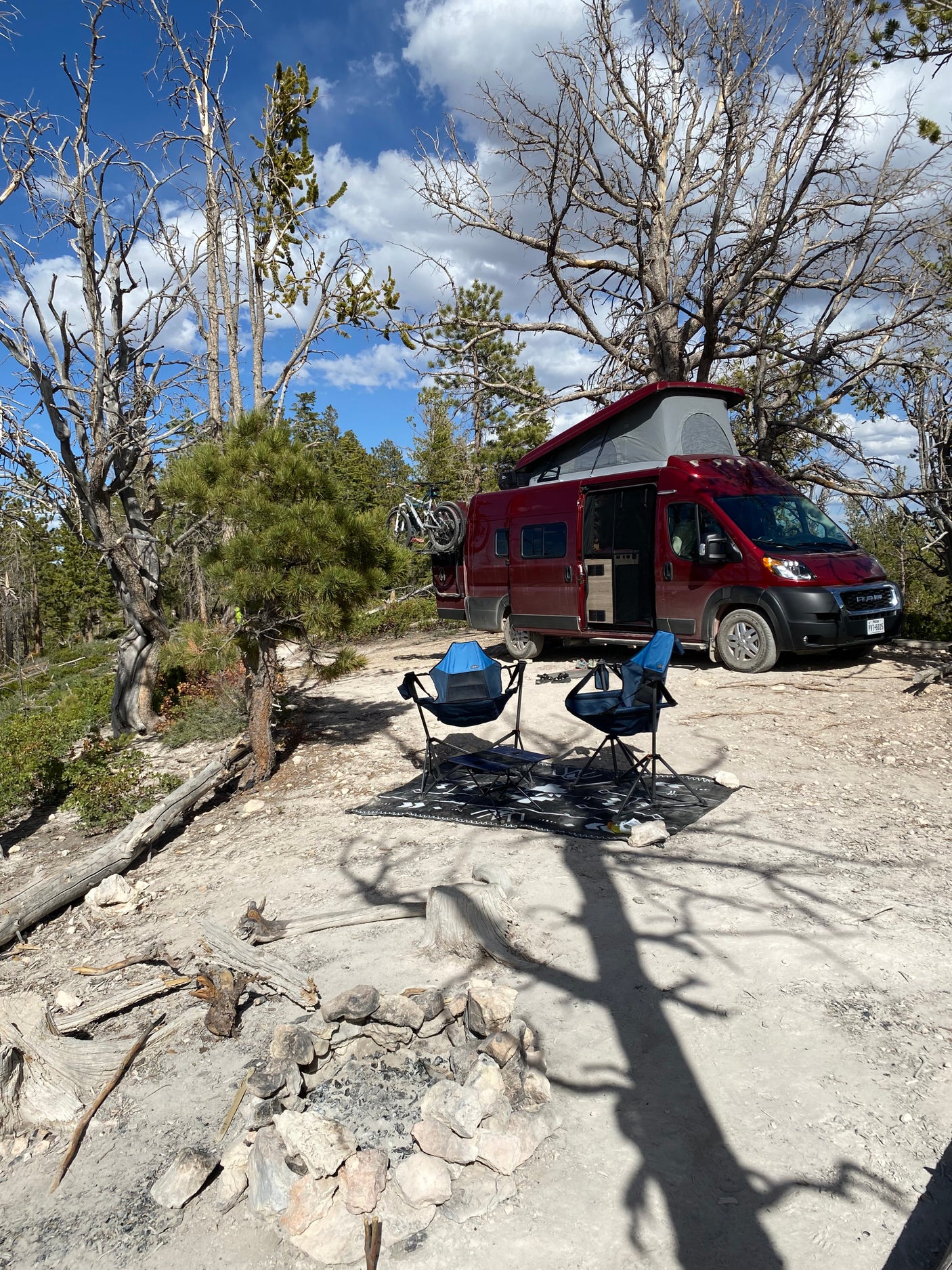 Scenic **rv rental** camping setup: Red van with bikes, chairs, & fire pit in a dry, forested wilderness area.