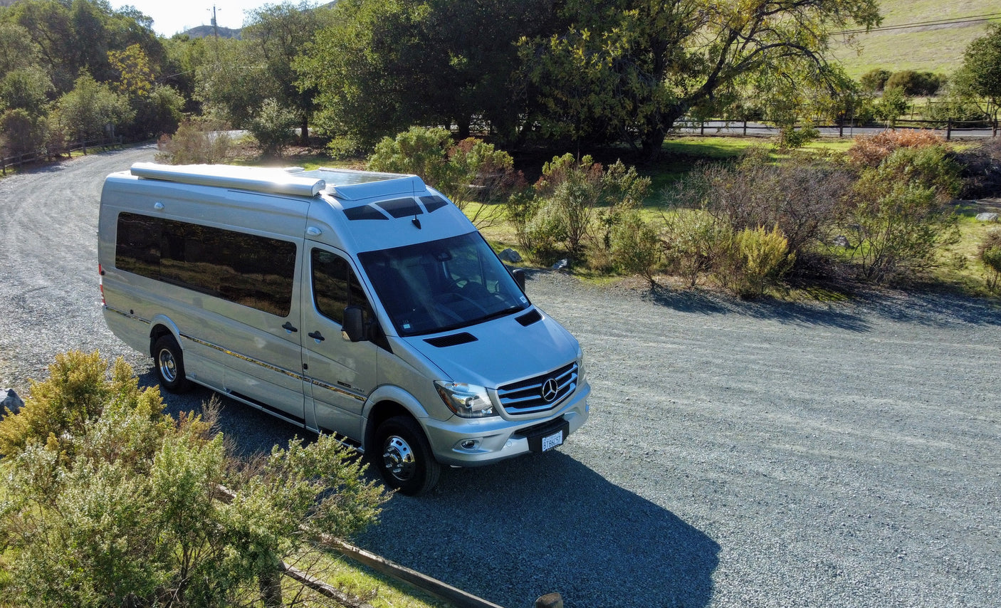 Silver Mercedes Sprinter RV rental, a sleek camper van, parked on a gravel road under sunny skies, surrounded by greenery.