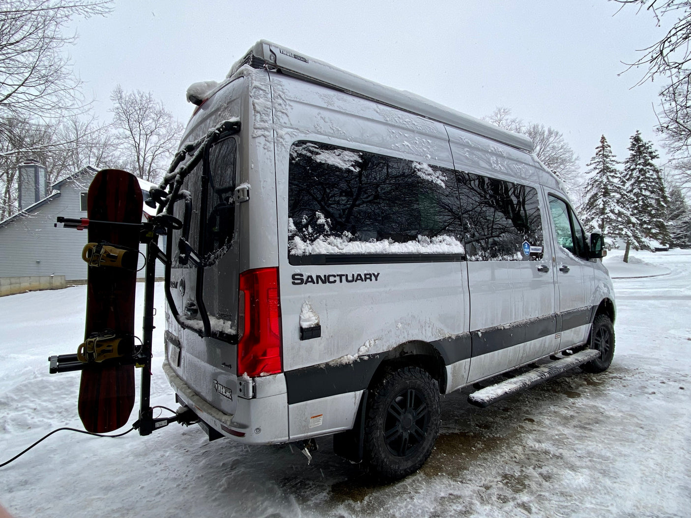 A snow-covered white RV rental camper van, with a snowboard mounted on the back, is parked in a snowy winter driveway.