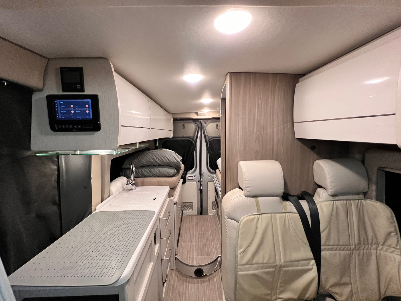 Inside an RV rental, a compact living space features a kitchen, bed area, passenger seats, and a wall control panel.