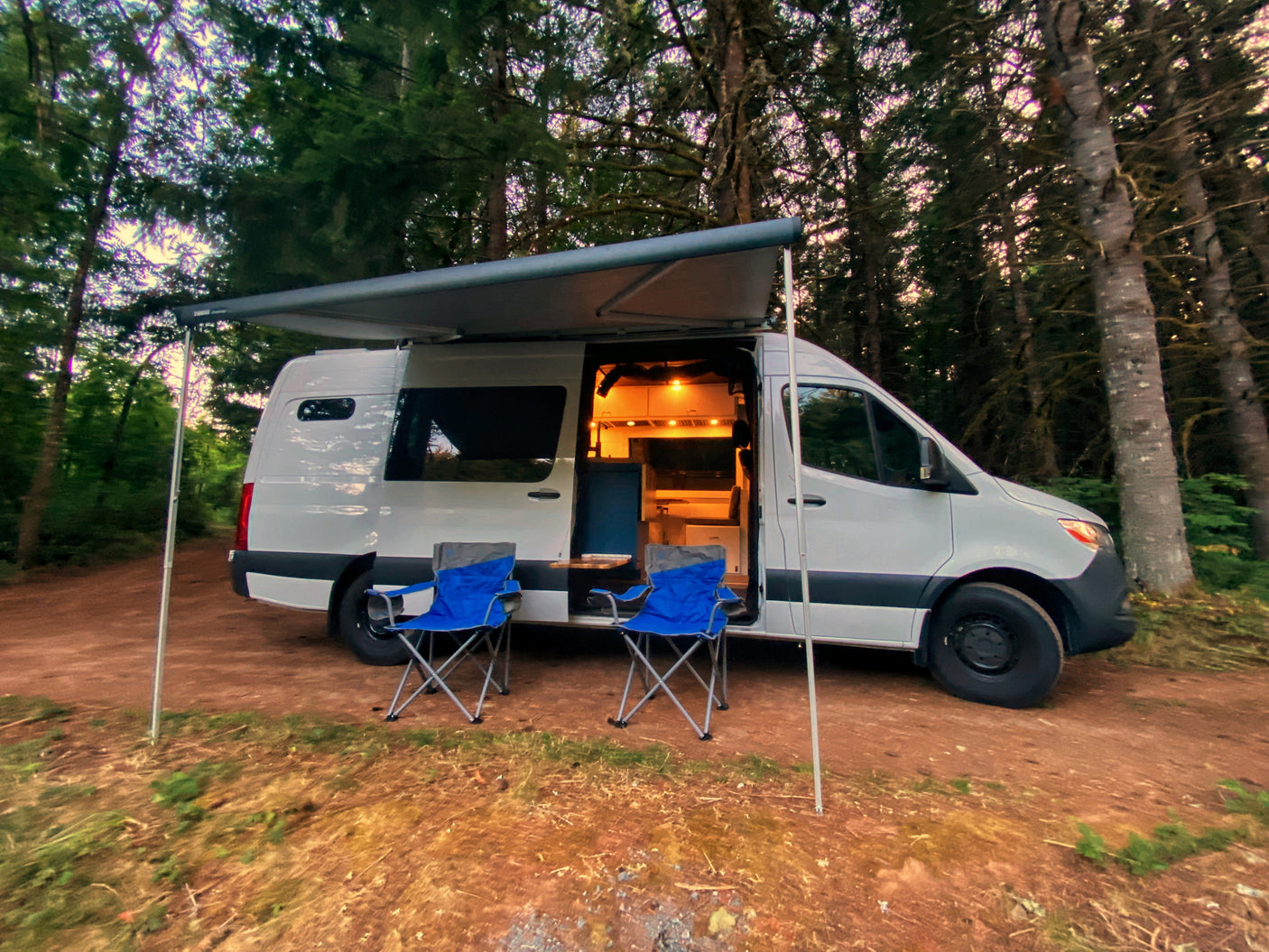 RV rental camper van in a forest with awning, lit interior, & chairs. Perfect for a cozy outdoor adventure.