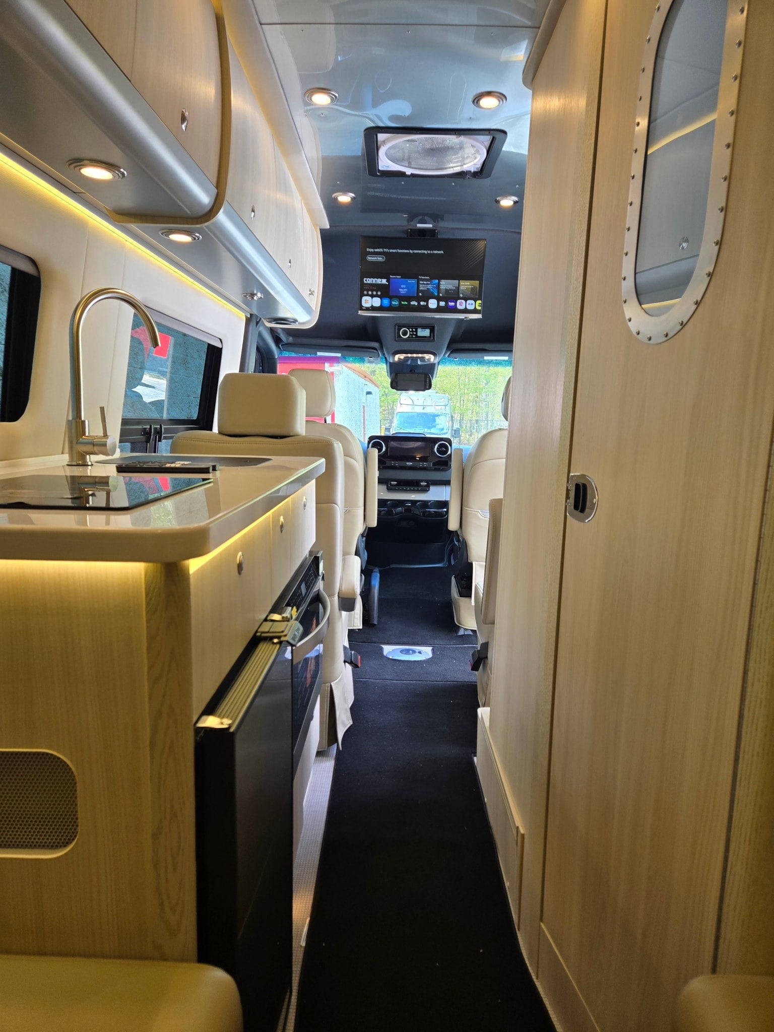Interior of an RV rental: a modern kitchen with a sink, cream seats, TV, and cockpit ahead, bathed in warm lighting.