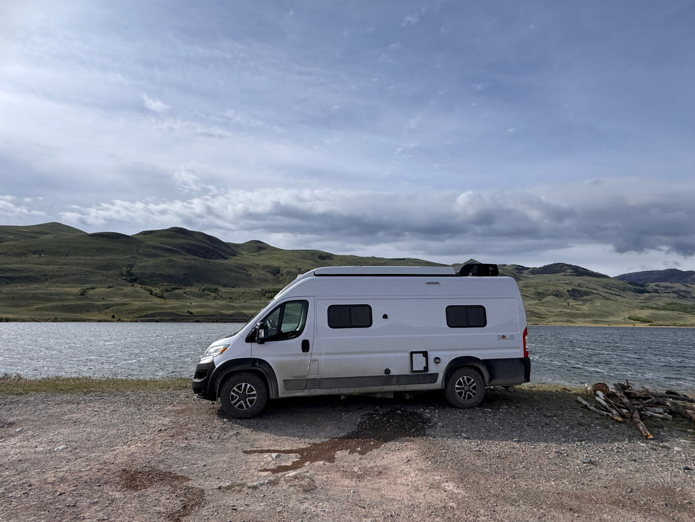 Winnebago Solis rv rental parked by a lake with rolling green hills & cloudy skies. Ready for adventure and scenic drives.