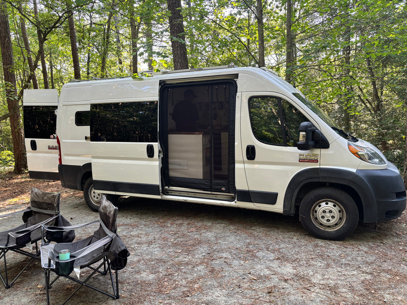 RV rental van, converted for camping, parked at a wooded site with chairs. Ready for adventure!