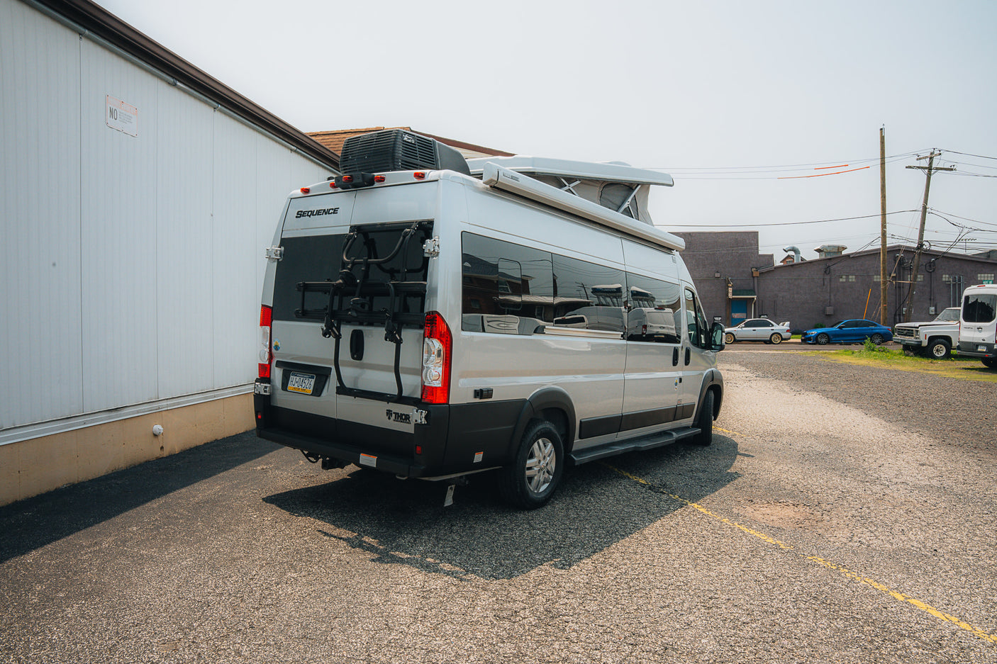 A light-colored Thor Sequence RV rental van, featuring a bike rack, parked on asphalt near a white building.