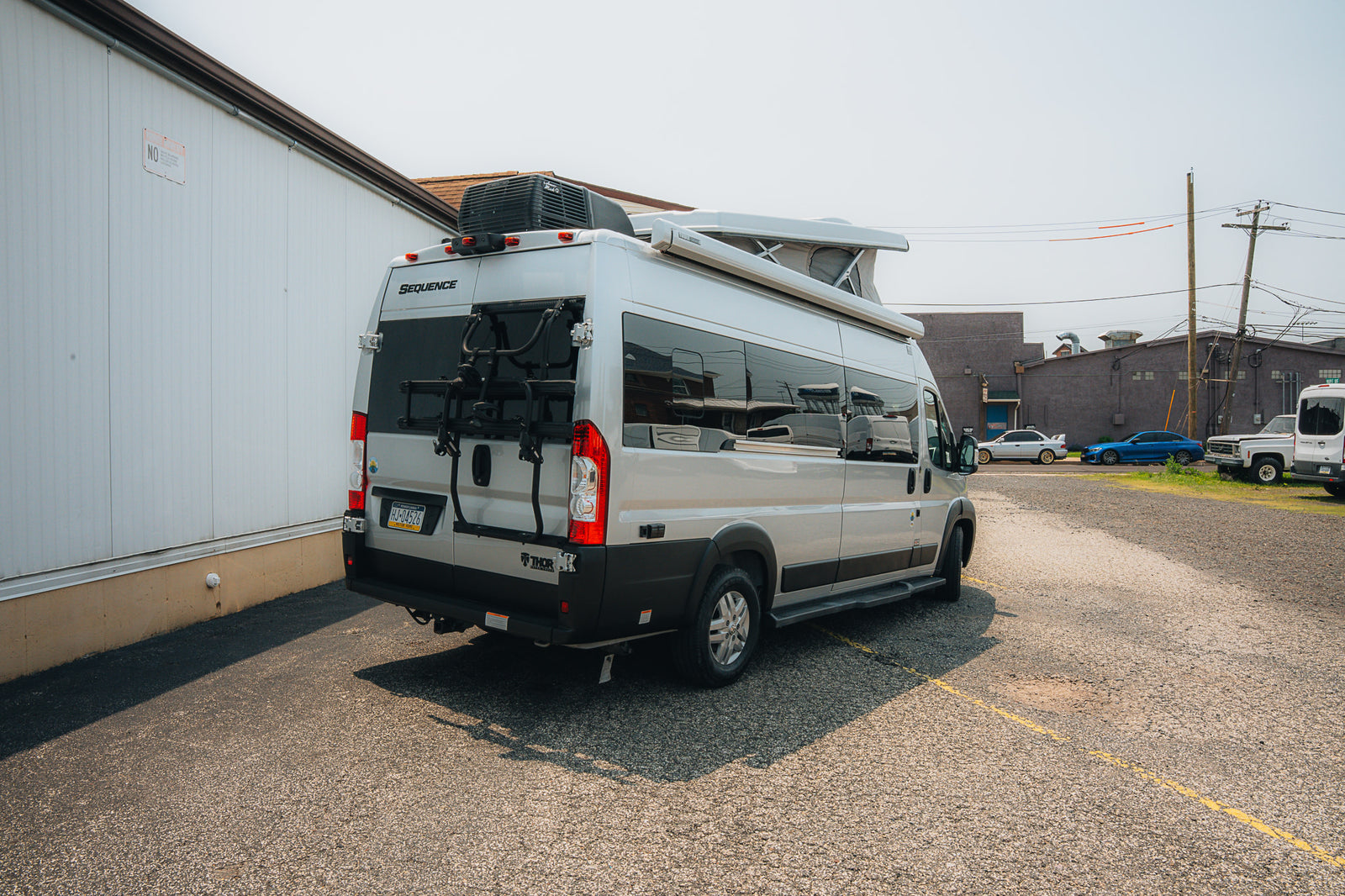 A light-colored Thor Sequence RV rental van, featuring a bike rack, parked on asphalt near a white building.