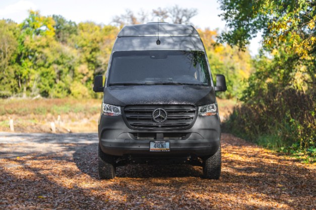 A dark grey Mercedes-Benz Sprinter camper van, marked '4RENT', on a leaf-covered path. Ideal for your next RV rental adventure.