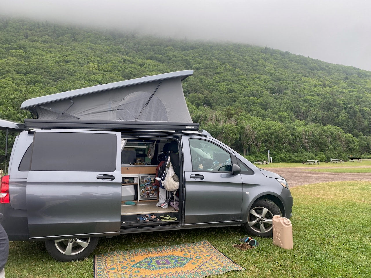 Grey pop-top campervan from an RV rental, parked for camping by a misty green mountain. Shows cozy van life setup.
