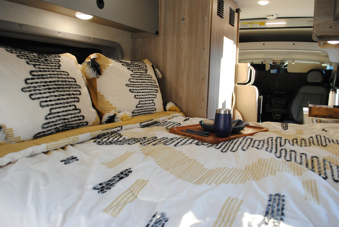 parked by a tranquil lake.|Silver Ram ProMaster RV rental by a calm lake with wooded hills. Ideal for outdoor adventures.|Inside a comfy RV rental: a bed with patterned bedding