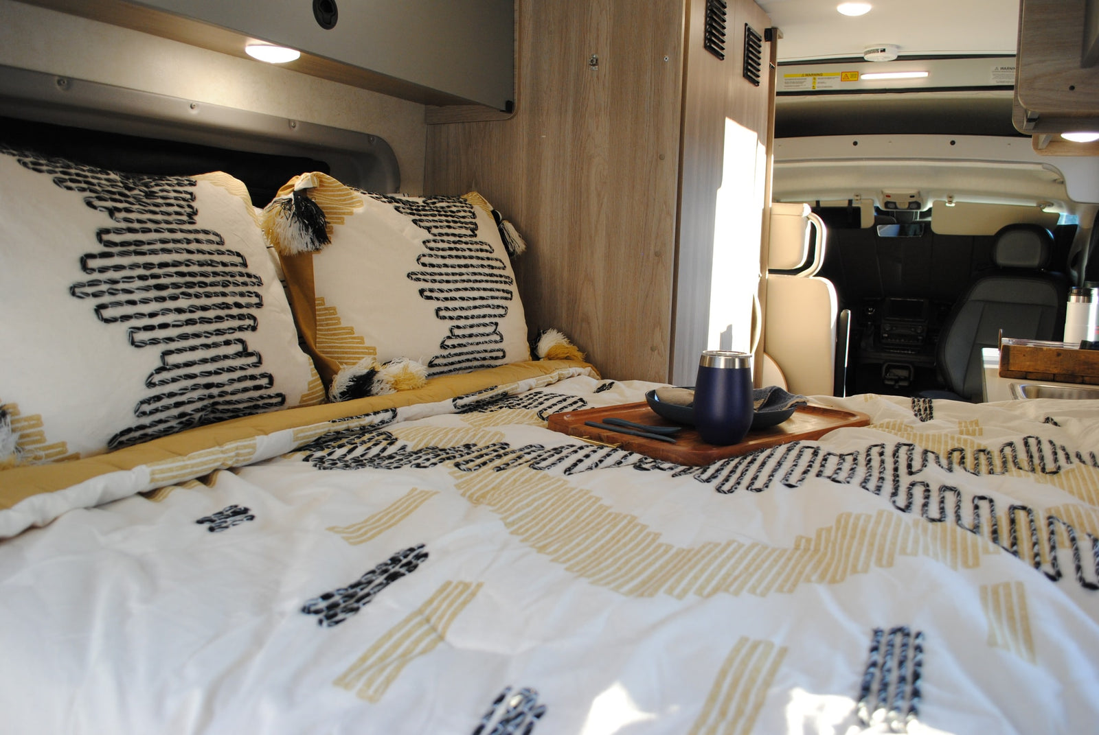 parked by a tranquil lake.|Silver Ram ProMaster RV rental by a calm lake with wooded hills. Ideal for outdoor adventures.|Inside a comfy RV rental: a bed with patterned bedding