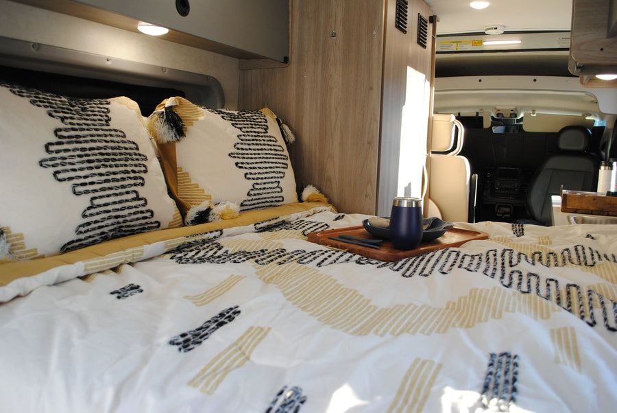 parked by a tranquil lake.|Silver Ram ProMaster RV rental by a calm lake with wooded hills. Ideal for outdoor adventures.|Inside a comfy RV rental: a bed with patterned bedding