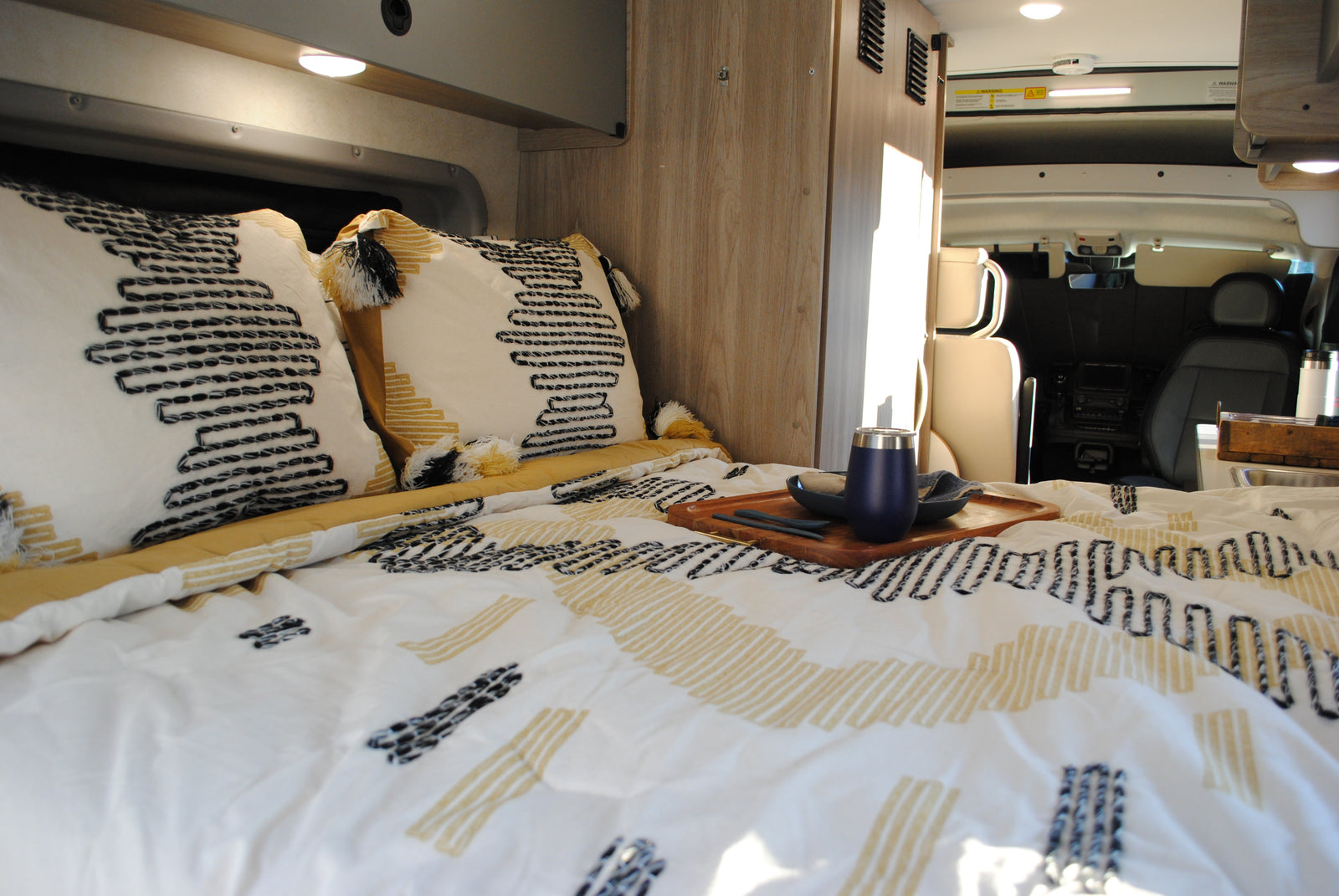 Inside a comfy RV rental: a bed with patterned bedding, decorative pillows, and a wooden serving tray. Front cab visible.