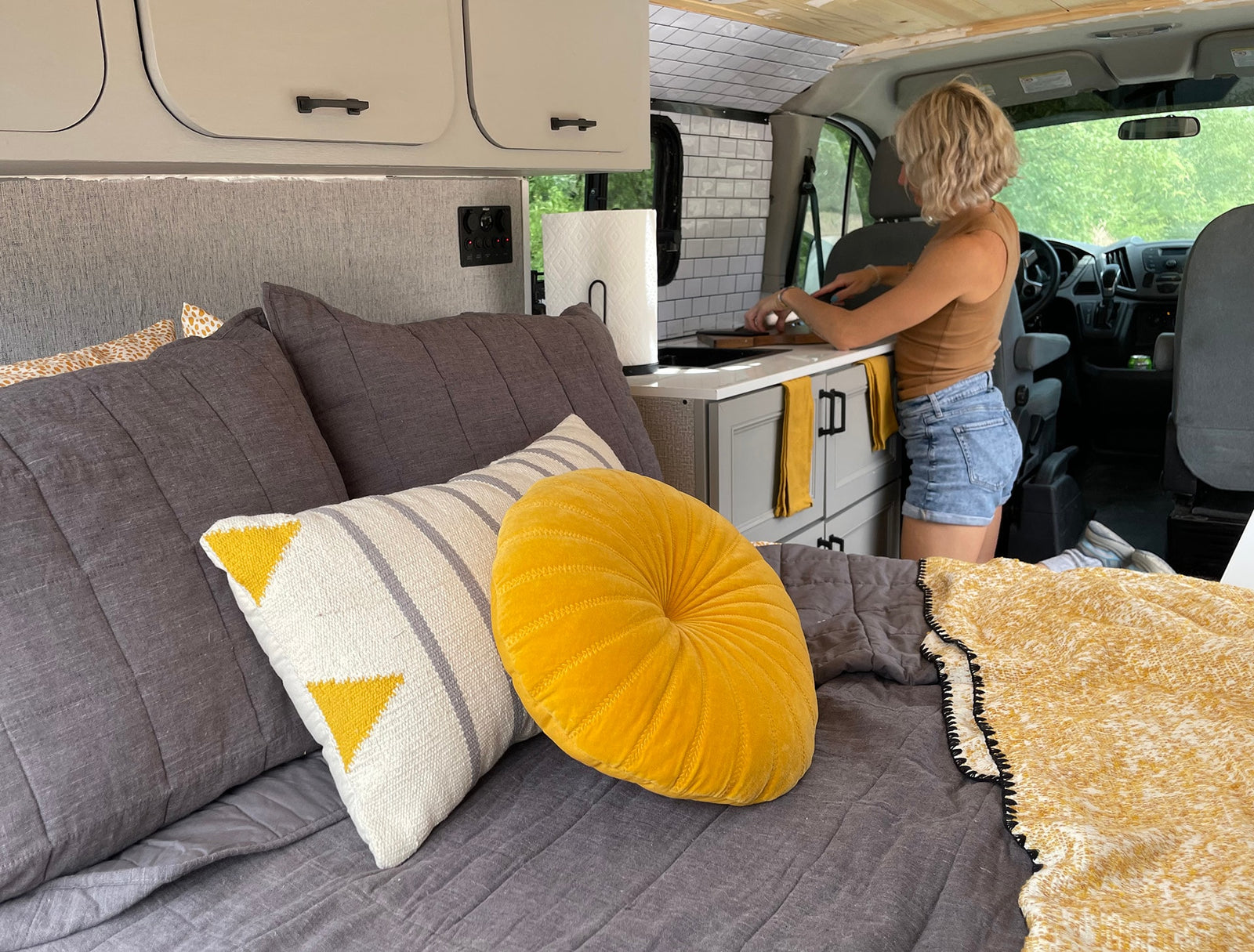 This RV rental shows a woman cooking in a compact kitchen, with a cozy bed featuring gray and yellow pillows in the foreground.