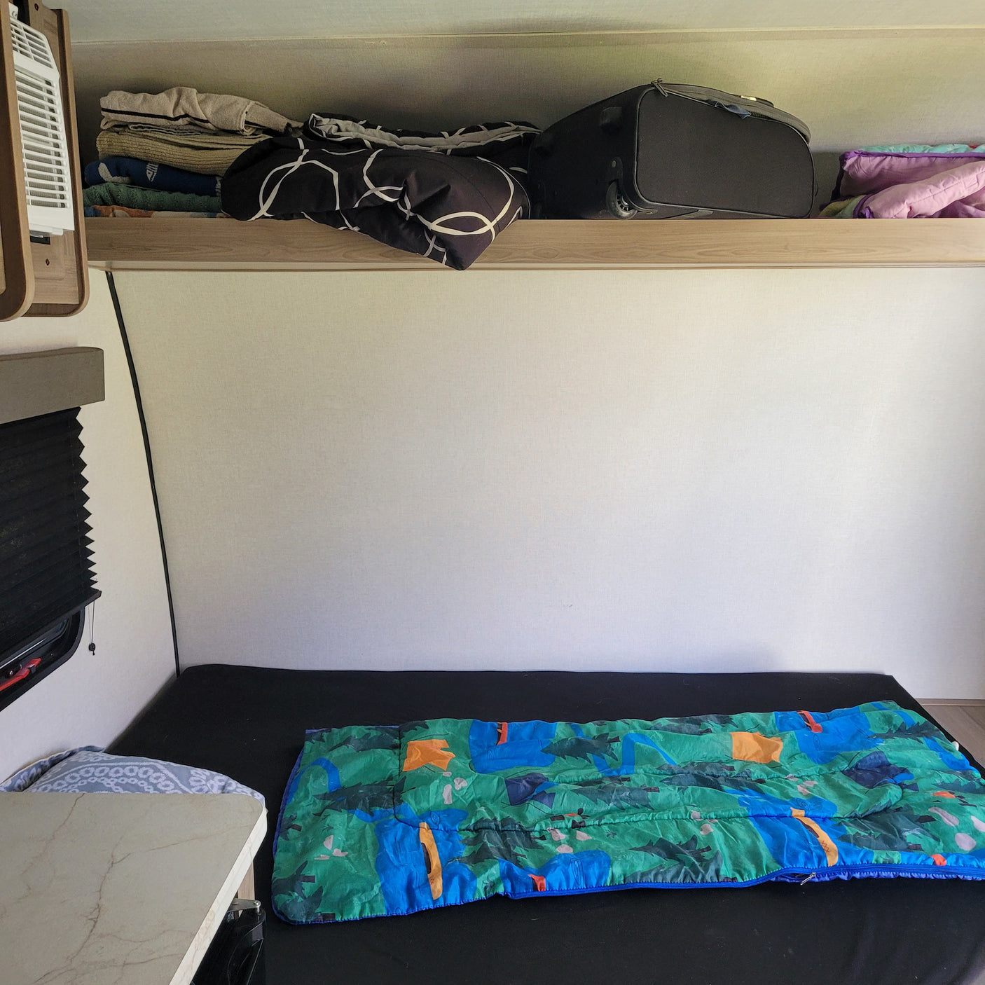 Cozy rv rental sleeping area with a colorful sleeping bag, overhead luggage, linen storage, and a small table.
