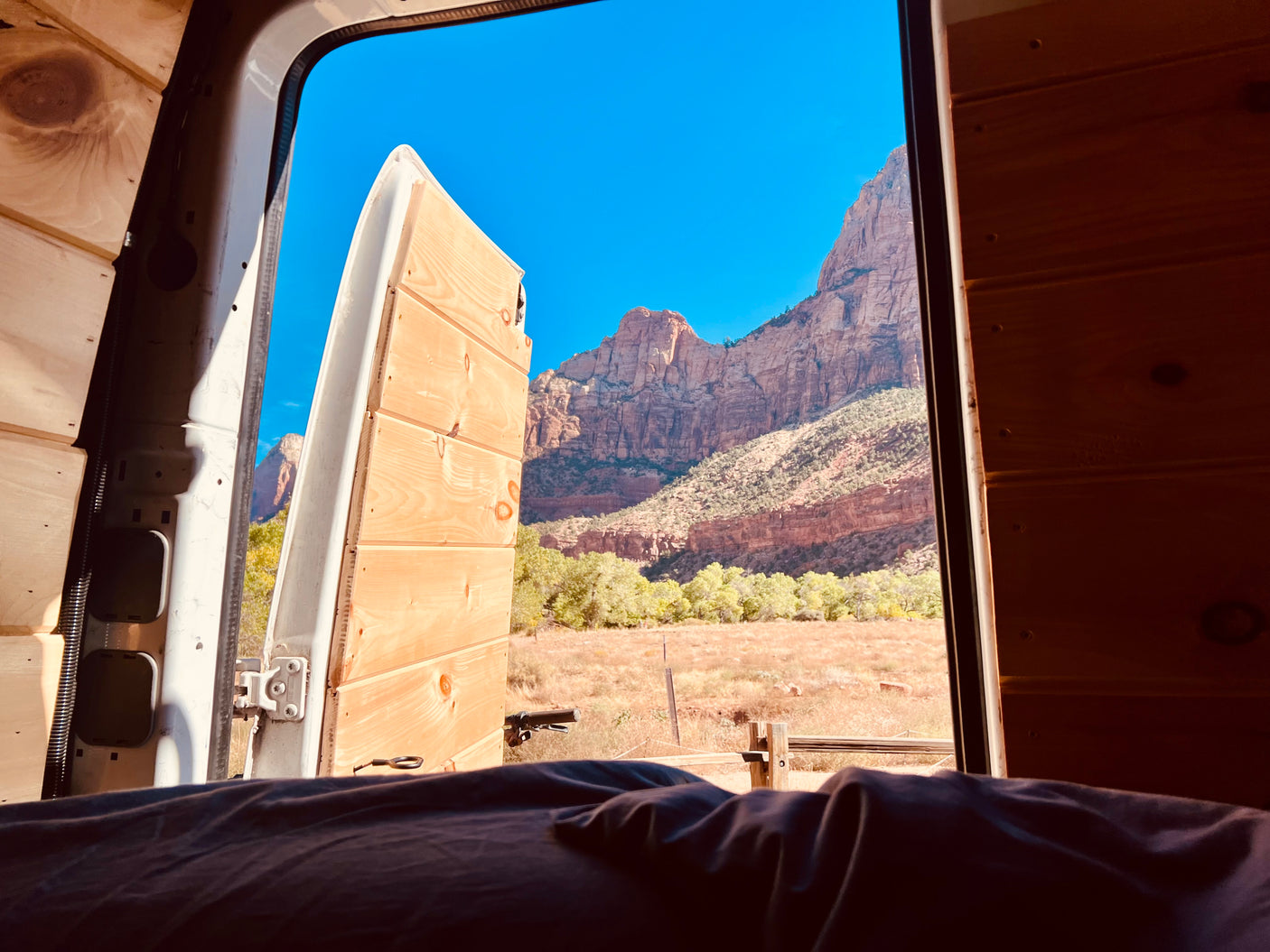 RV rental view: Majestic red mountains, green trees, & field through open doors of a cozy, wood-paneled van.