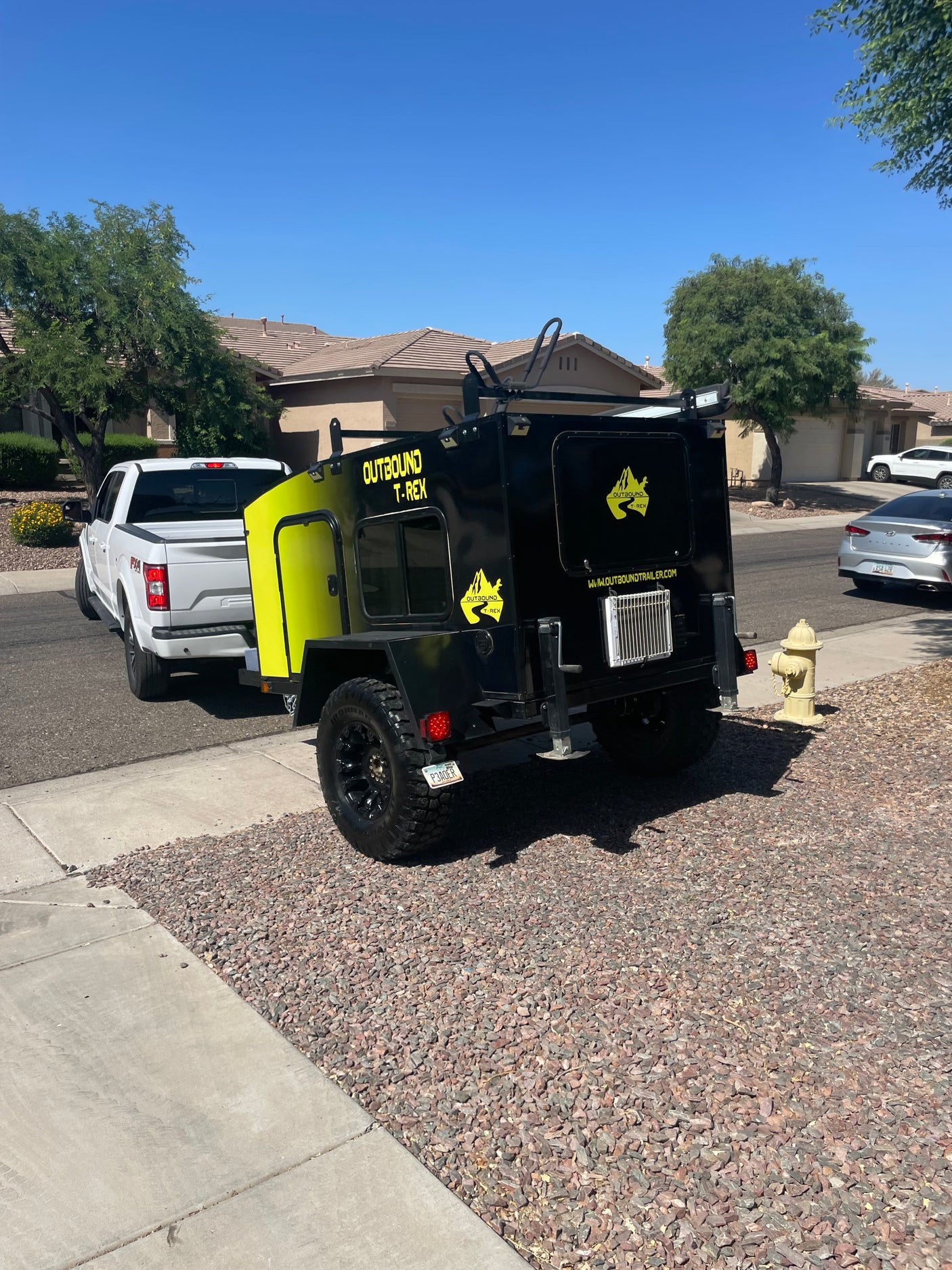 An Outbound T-REX off-road travel trailer for RV rental, hitched to a white pickup truck on a sunny day.