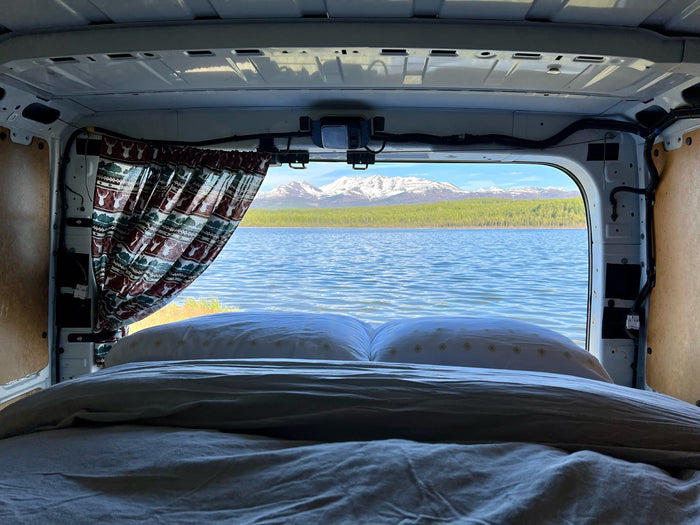 cooler. Perfect for outdoor adventures.|RV rental interior: A made bed with a patterned blanket looks out to a lake view through open rear doors.|Scenic RV rental view: Cozy bed overlooking a calm lake and snow-capped mountains. Perfect for van life adventures!|White Nissan van RV rental parked by an Alaskan lake