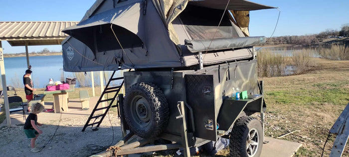 set up at a lakeside campsite.|Rugged off-road trailer with a Nomadic Extended rooftop tent setup at a campsite. An alternative to an RV rental for camping.|An adventure-ready rv rental: a rugged camping trailer with a rooftop tent