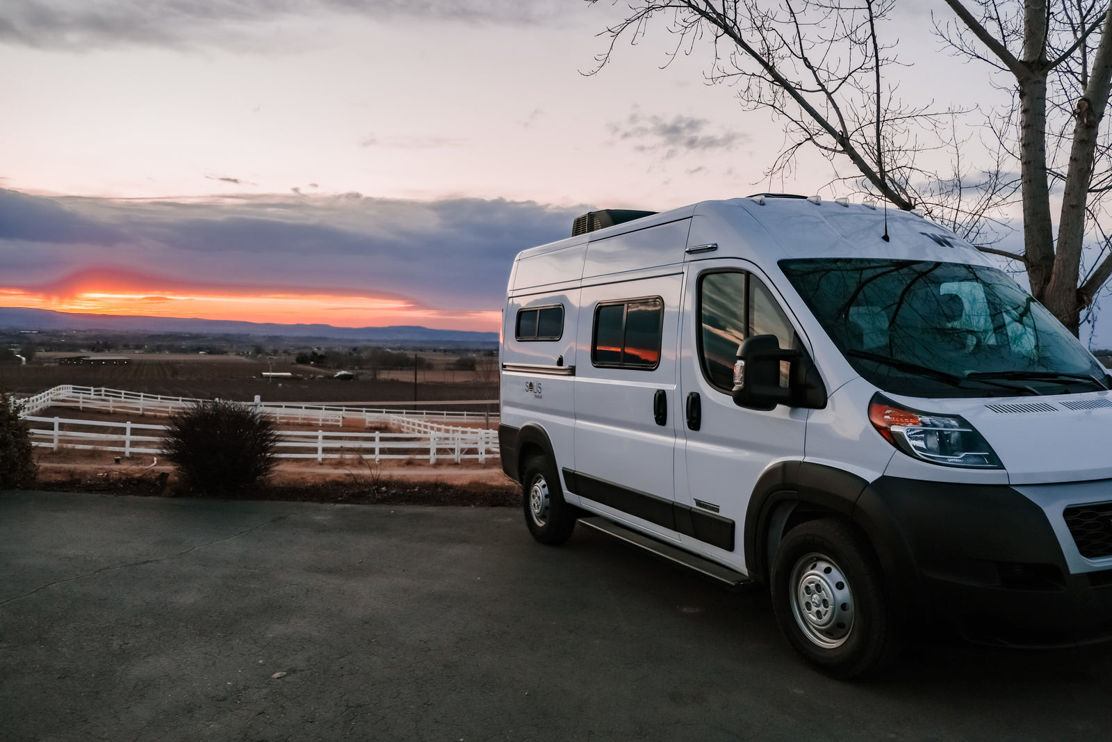 White Winnebago Solis Pocket RV rental at sunset, parked with a vibrant orange sky over a fenced valley.