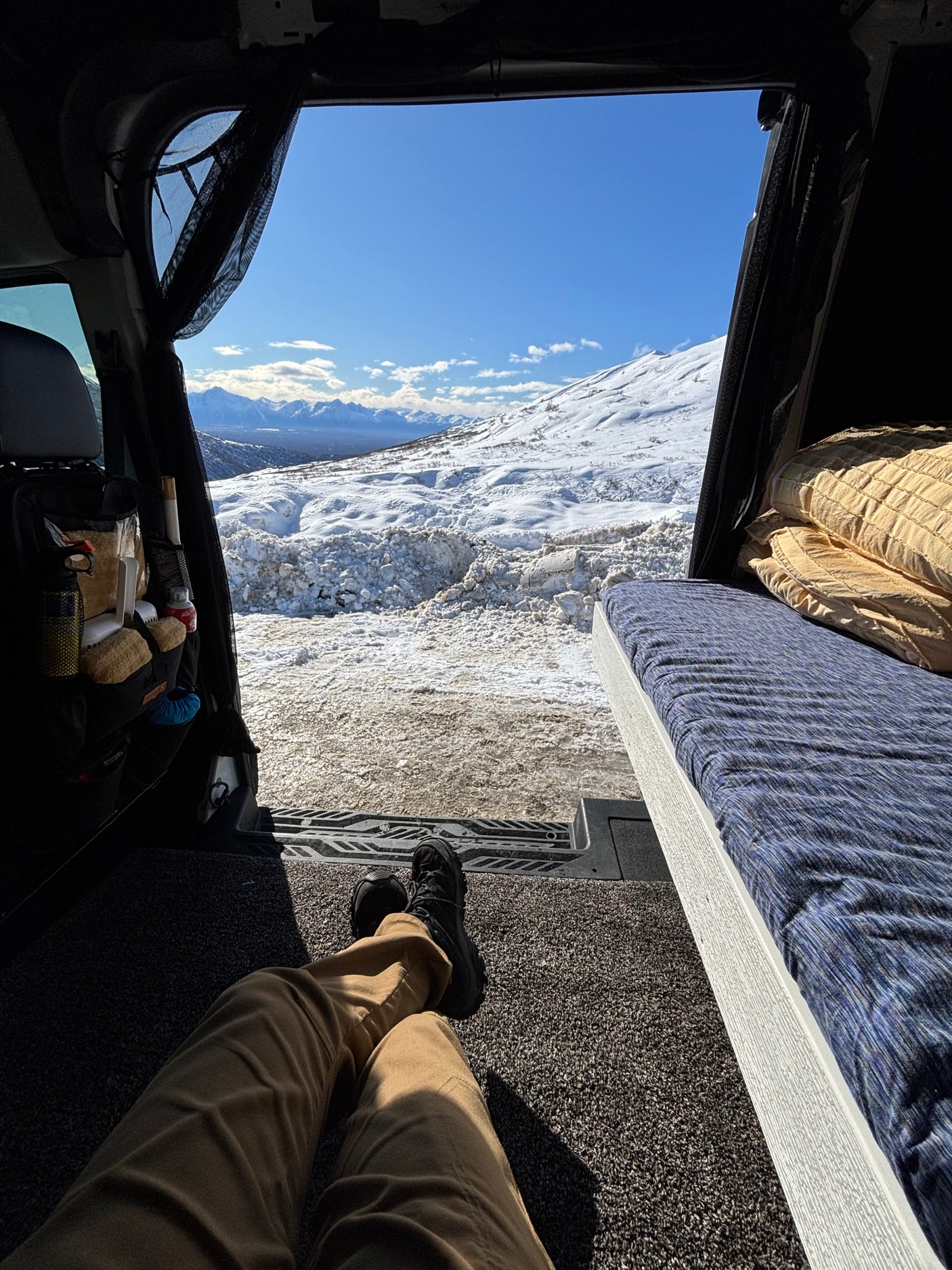View from an RV rental's rear, showing a person's legs, interior bed, and a breathtaking snowy mountain landscape.