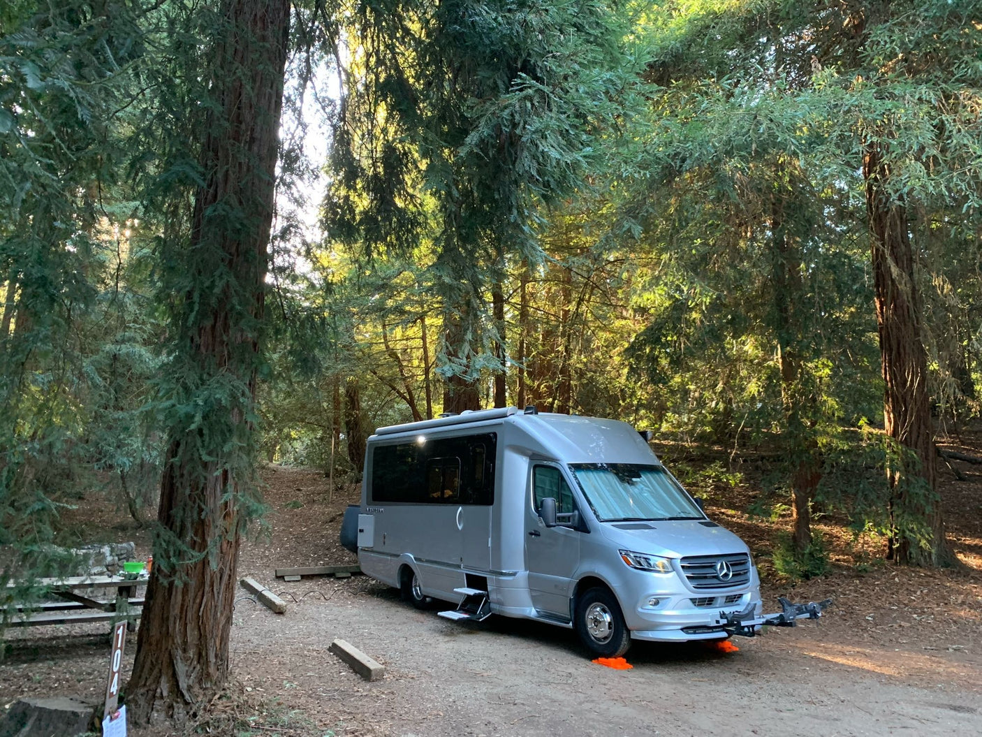 Silver RV rental settled at campsite 104 among towering trees. Ideal for a serene forest getaway, with a picnic area and bike rack.