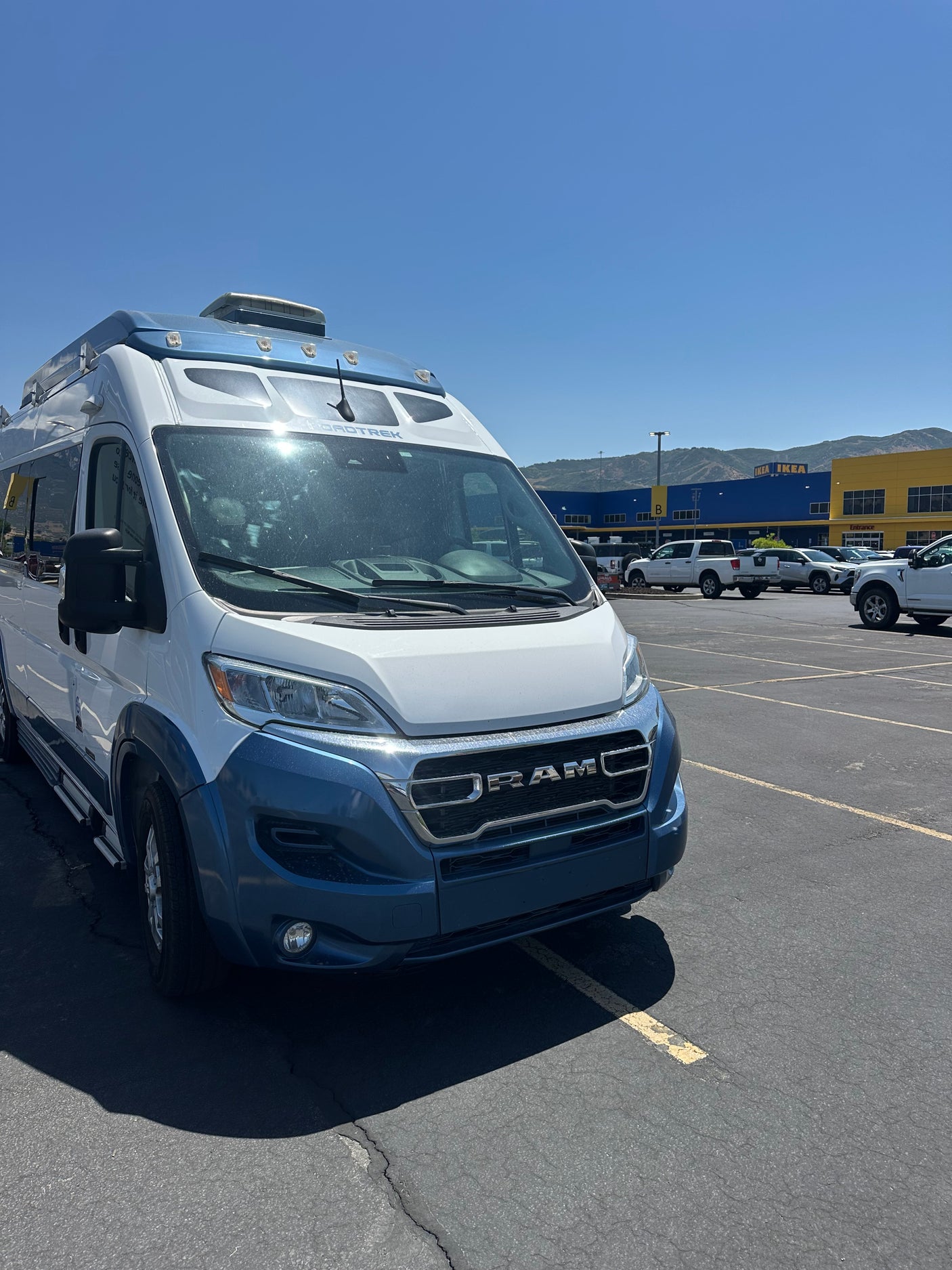 Roadtrek RV rental (white/blue) in a sunny parking lot, with an IKEA store and mountains in the distance.