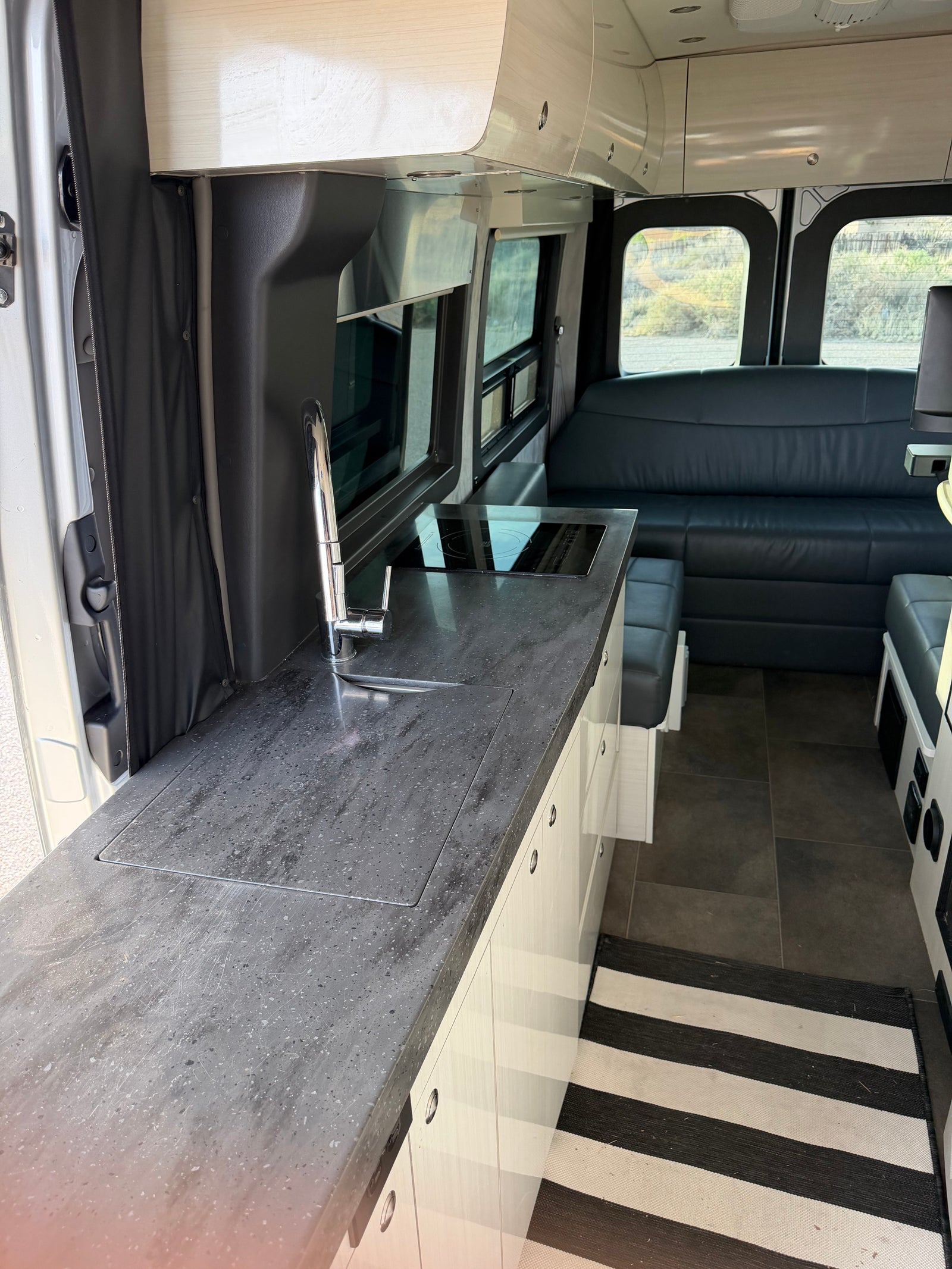 Modern rv rental interior: kitchenette (sink, induction cooktop), sleek white cabinets, dark floor, striped rug, and rear seating.