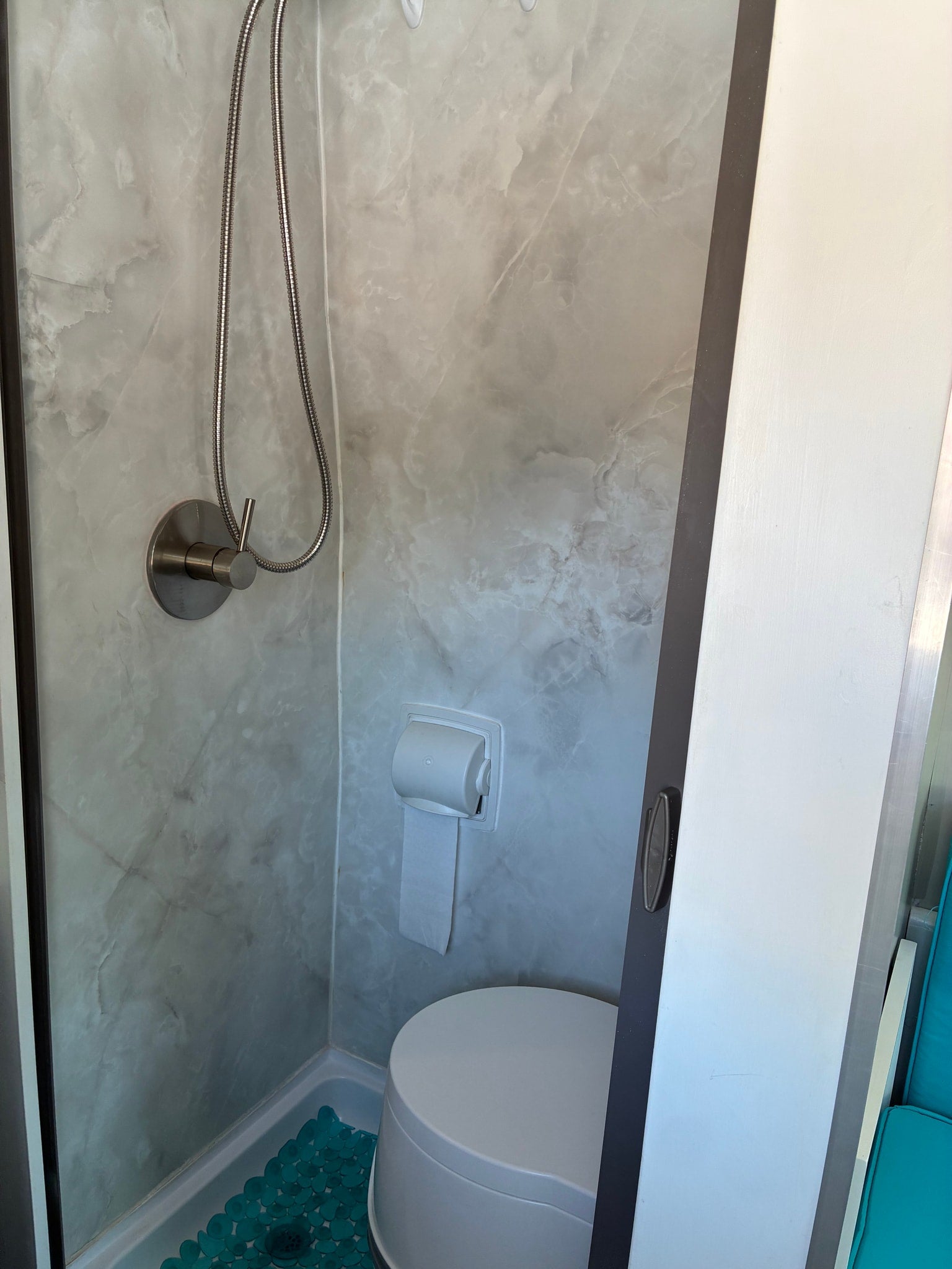 RV rental compact bathroom with shower, toilet, toilet paper, grey marble walls, and a teal floor mat.
