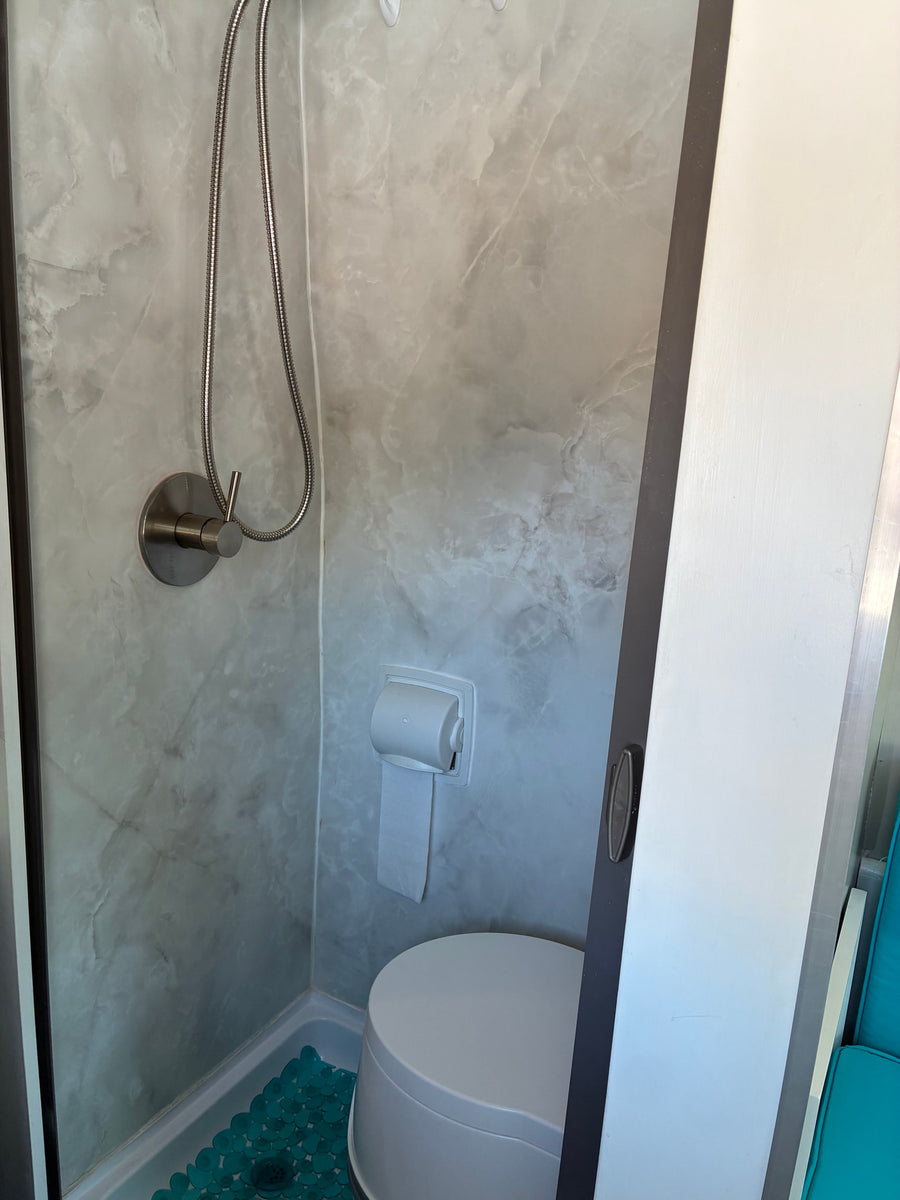 RV rental compact bathroom with shower, toilet, toilet paper, grey marble walls, and a teal floor mat.