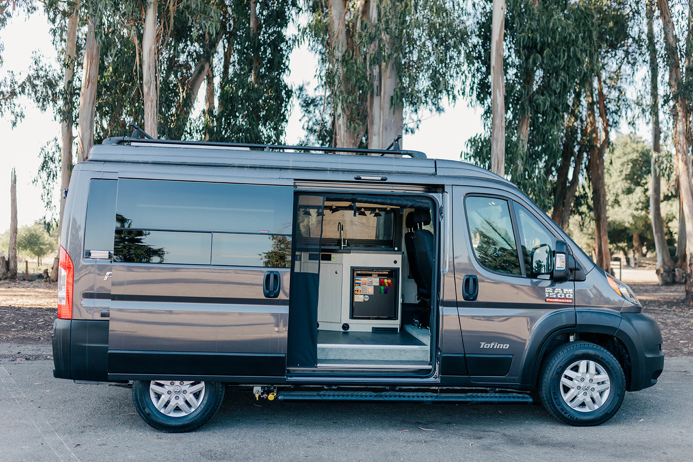 Gray Ram ProMaster camper van RV rental, side door open to reveal a compact kitchen & fridge. Perfect for adventure.