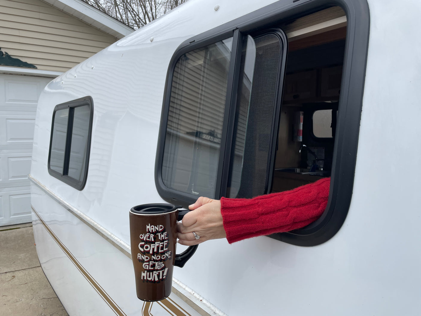 An arm in a red sweater holds a "Hand over the coffee" mug out an RV rental window.
