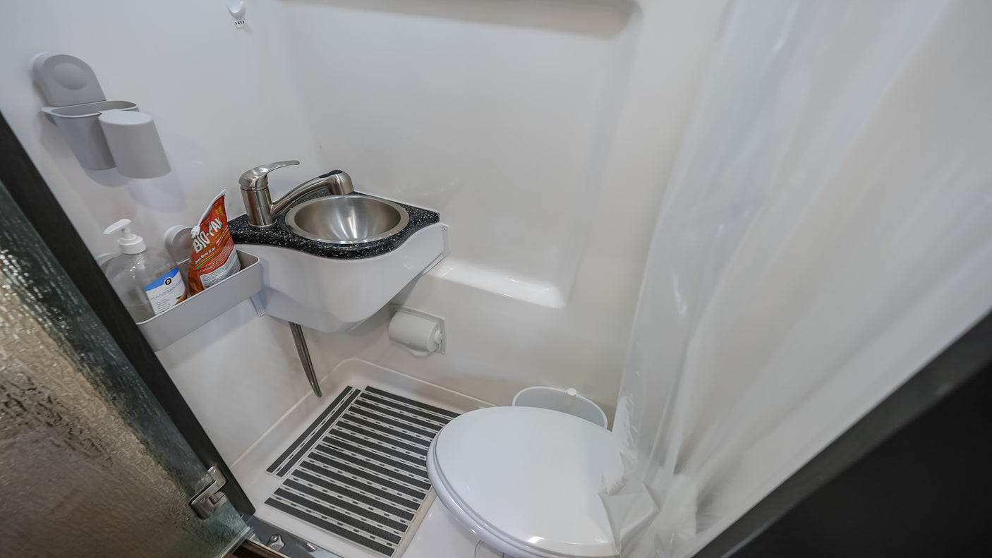 Inside an RV rental: a compact bathroom with toilet, sink, shower, hand sanitizer, and floor mat for comfort.