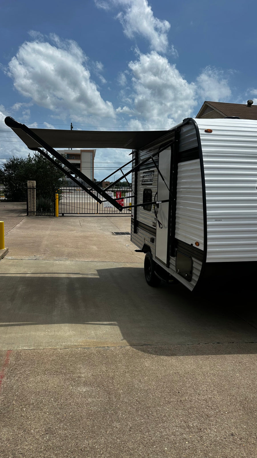 A white rv rental travel trailer with its dark awning extended on a sunny paved lot under a blue, cloudy sky.