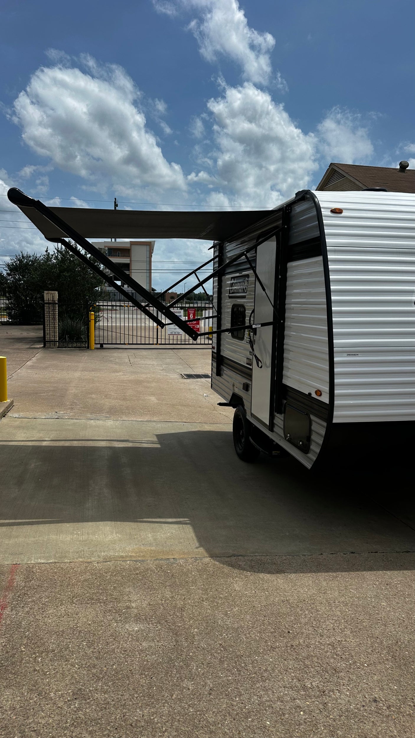 A white rv rental travel trailer with its dark awning extended on a sunny paved lot under a blue, cloudy sky.