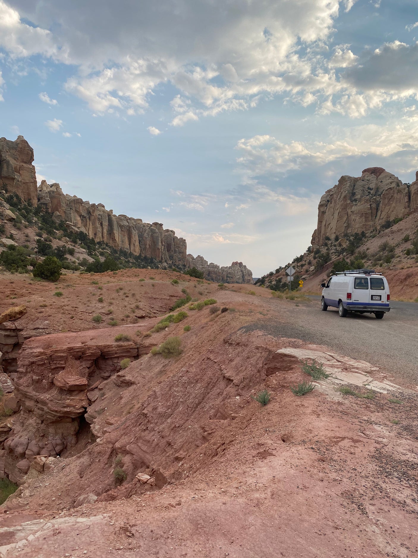 White van, possibly an RV rental, on a winding desert road within a canyon of towering red rock cliffs under cloudy skies.