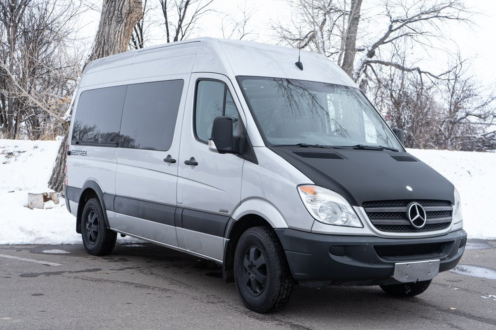 Silver Mercedes Sprinter van, an RV rental with a black hood, parked in snowy terrain. Your adventure camper awaits!