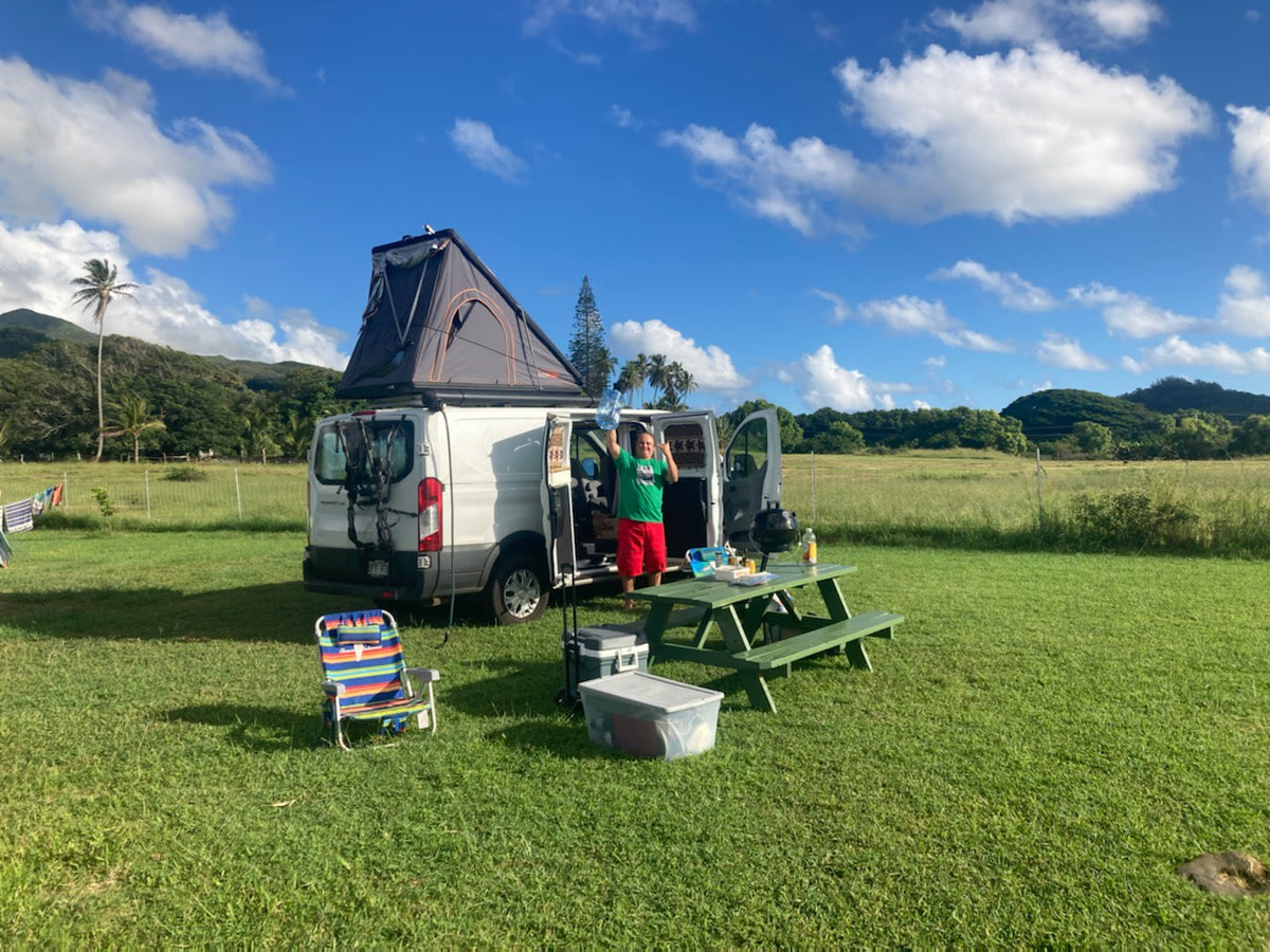 Enjoying van life with an RV rental: A white van with a rooftop tent and a happy person by a picnic table in a grassy field.|White Ford Transit campervan