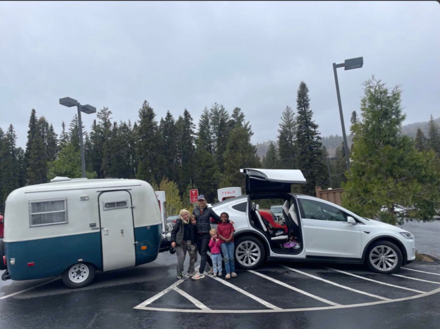 A family and their white Tesla Model X with a retro RV rental are seen charging at a Tesla Supercharger station. Road trip ready!